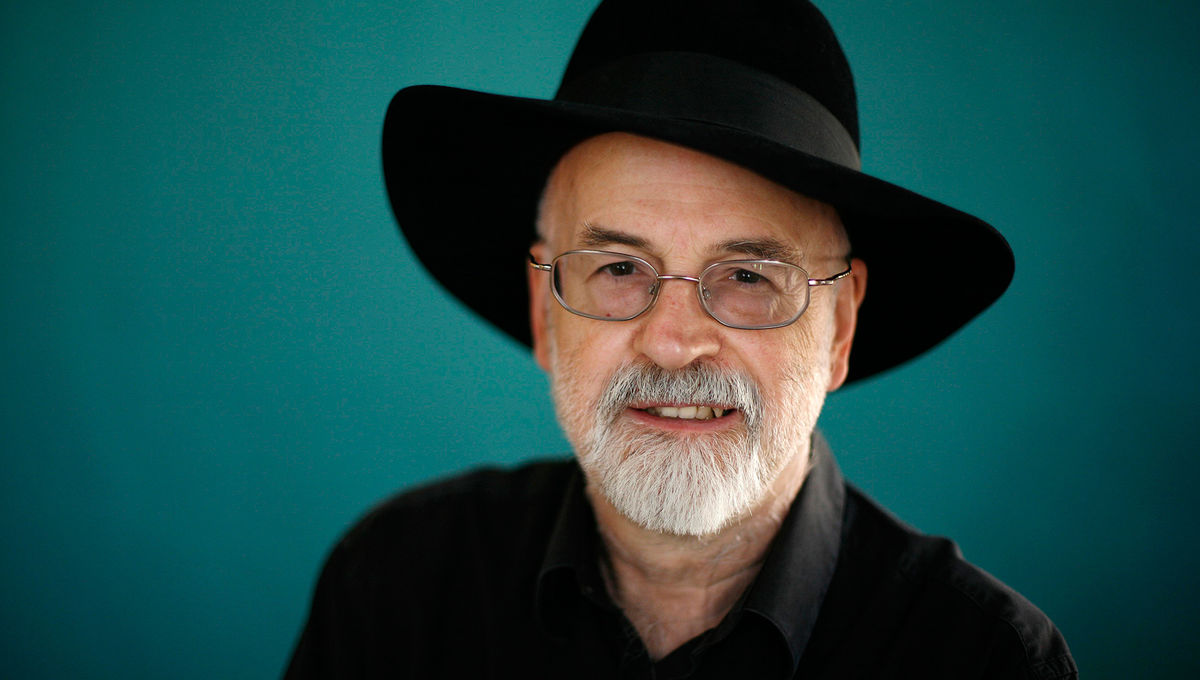 A headshot of Sir Terry Pratchett, wearing one of his infamous black hats, stood in front of a turquoise background.